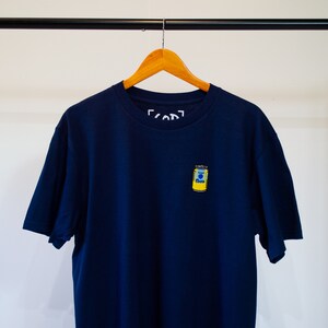 Op de afbeelding: Een donkerblauw T-shirt met een geel en blauw blikontwerp op de voorkant. Het blik heeft het woord "Soda" erop.