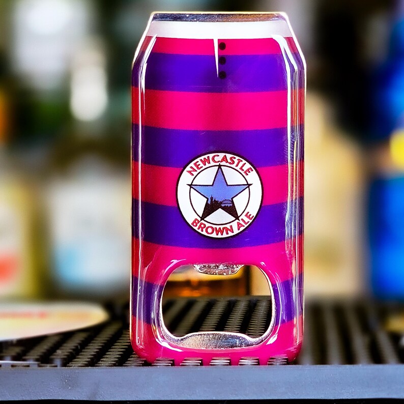 May include: A bottle opener designed to look like a can of Newcastle Brown Ale. The opener has a pink and purple striped pattern with the Newcastle Brown Ale logo. The opener is made of metal and plastic.