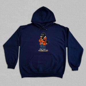 Puede incluir: Sudadera con capucha azul marino con un personaje de dibujos animados de un p&aacute;jaro que lleva gafas de sol, una chaqueta naranja y vaqueros. El p&aacute;jaro tiene los brazos cruzados. La sudadera tiene un bolsillo delantero y una capucha con cord&oacute;n.