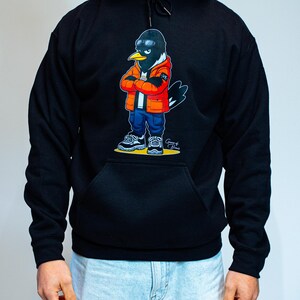 Puede incluir: Sudadera con capucha negra con un personaje de dibujos animados de un p&aacute;jaro que lleva un pasamonta&ntilde;as negro, una chaqueta naranja, vaqueros azules y zapatillas deportivas. El p&aacute;jaro tiene los brazos cruzados y lleva gafas de sol. El dise&ntilde;o est&aacute; impreso en la parte delantera.