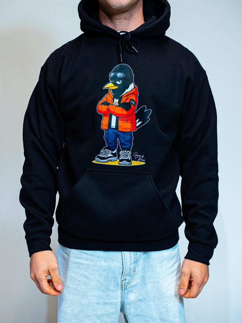 Puede incluir: Sudadera con capucha negra con un personaje de dibujos animados de un p&aacute;jaro que lleva un pasamonta&ntilde;as negro, una chaqueta naranja, vaqueros azules y zapatillas deportivas. El p&aacute;jaro tiene los brazos cruzados y lleva gafas de sol. El dise&ntilde;o est&aacute; impreso en la parte delantera.