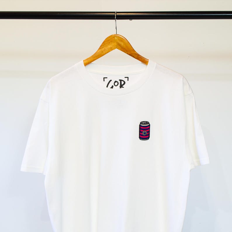 K&ouml;nnte beinhalten: Wei&szlig;es T-Shirt mit einem Design einer rosa-schwarzen gestreiften Dose und dem Text "[COR]" auf der Vorderseite.