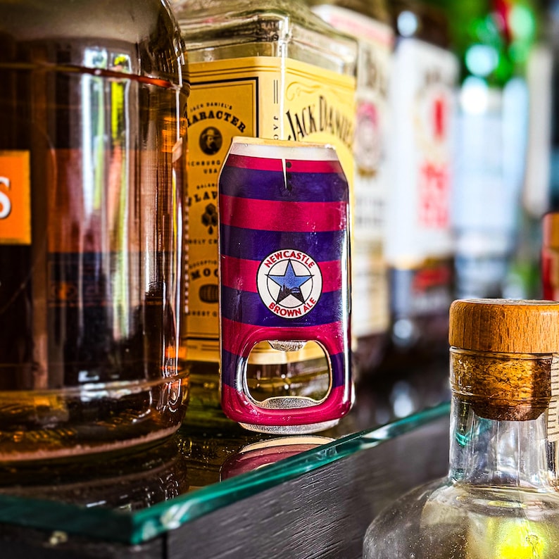 May include: A bottle opener designed to look like a Newcastle Brown Ale can. The opener is painted with alternating red and purple stripes and features the Newcastle Brown Ale logo. The opener is made of metal and has a bottle cap opener.