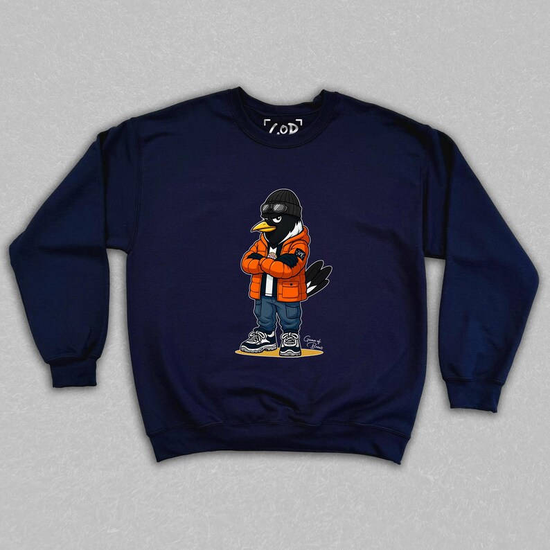 May include: Navy blue sweatshirt featuring a cartoon raven character wearing an orange jacket, black beanie, and jeans. The raven has its arms crossed and is wearing sunglasses. The sweatshirt has the text "/.OD" on the top.