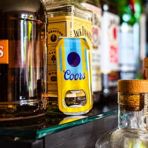 Pode incluir: Um abridor de garrafa de cerveja Coors amarelo e azul com um orifício circular para abrir garrafas. O abridor tem um círculo azul e listras azuis verticais. O fundo inclui garrafas de licor.