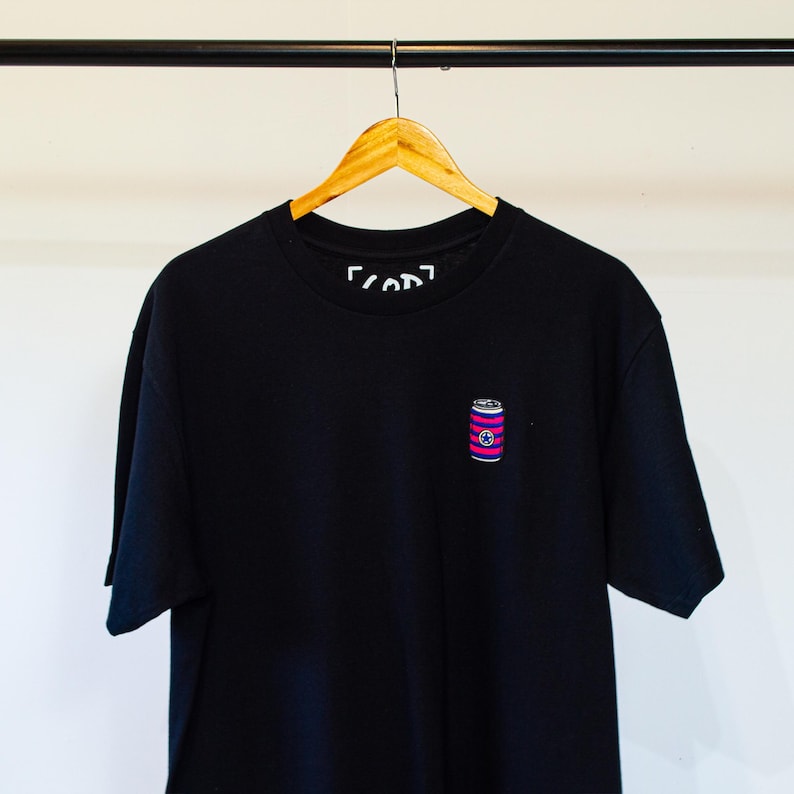 K&ouml;nnte beinhalten: Schwarzes T-Shirt mit einem kleinen gestickten Bild einer rosa und blauen Dose mit einem wei&szlig;en Stern. Der Text "[COD]" ist am Kragen sichtbar.