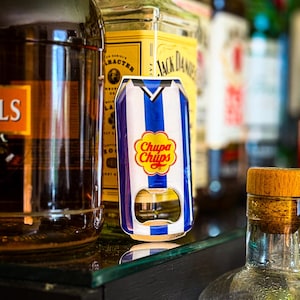 May include: A Chupa Chups-branded bottle opener in the shape of a soda can, featuring blue and white stripes and the Chupa Chups logo. The opener is displayed on a glass surface, with various liquor bottles in the background.