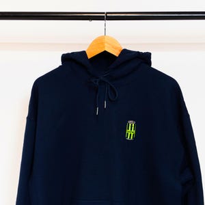以下が含まれることがあります： 前面に緑と黒の缶のグラフィックがプリントされたネイビーブルーのパーカー。缶には「SOCO」という文字が印刷されています。