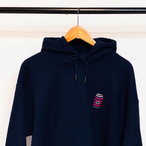 May include: A navy blue hoodie with a red and white striped can design on the front. The can has a red circle with a white star inside.