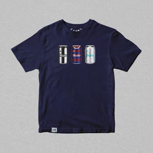 K&ouml;nnte beinhalten: Ein marineblaues T-Shirt mit einem Aufdruck von drei Aluminiumdosen. Jede Dose ist mit dem Trikot eines anderen Fu&szlig;ballteams gestaltet. Die Dosen sind mit "Basela" beschriftet.