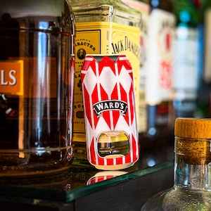 May include: A red and white bottle opener with the text "WARD'S" in a black banner. The opener is shaped like a can and is sitting on a glass shelf. Several bottles of liquor are in the background.