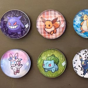 Könnte beinhalten: Zwölf runde Magnete mit verschiedenen Pokémon-Charakteren. Die Magnete sind bunt und haben eine glänzende Oberfläche.