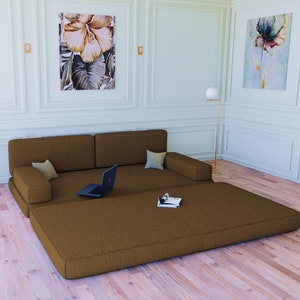 May include: A brown futon sofa with a brown mattress unfolded on the floor. The sofa has two pillows and a laptop on the seat. The room has white walls and wood floors.