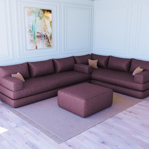 Puede incluir: Un gran sofá de esquina marrón con un puf a juego. El sofá tiene varios cojines y está colocado en una esquina de una habitación con paredes blancas y un cuadro en la pared.