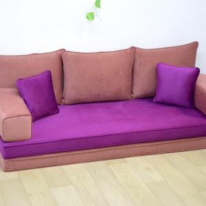 Puede incluir: Un sofá de suelo rosa y morado con tres almohadas. El sofá tiene una base rosa y una parte superior morada con un reposabrazos rosa a cada lado. Las almohadas son rosas y moradas.