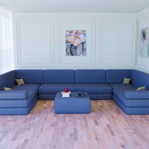 Könnte beinhalten: Ein großes, modulares blaues Sofa mit passendem Hocker in einem hellen Raum. Das Sofa ist U-förmig angeordnet, mit mehreren gelben Kissen. Ein kleiner Tisch enthält einen Laptop und einen roten Apfel. Der Raum hat hellfarbene Wände und Holzböden.