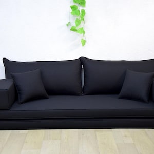 以下が含まれることがあります： 3つのクッションと1つのアームレストが付いた黒い布団ソファ。ソファは折り畳まれており、使用できます。ソファは木製の床の上にあります。