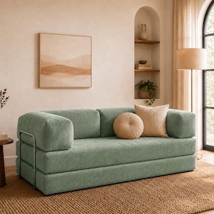 May include: A sage green corduroy sofa featuring a low-profile design with thick, blocky armrests and a metal frame support. The sofa is accented with one round beige cushion and one square beige cushion, providing a textured seating surface.