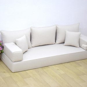 May include: A white floor sofa with a thick cushion and four white pillows. The sofa is sitting on a light wood floor. The text "FloorSeatings" is visible on the sofa.