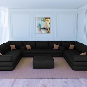 May include: A large black sectional sofa with a matching black ottoman. The sofa has multiple cushions and is arranged in a U-shape. The sofa is in a room with white walls and a painting on the wall.