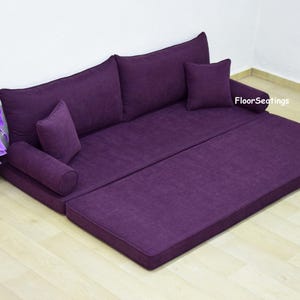 Op de afbeelding: Een paarse vloerzitbank met drie kussens en een rugleuning. De bank is gemaakt van een zachte, pluche stof en is perfect om te loungen of te slapen. De bank staat op een lichte houten vloer.