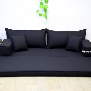 Puede incluir: Un sofá de suelo negro con dos cojines grandes de respaldo, dos cojines más pequeños y dos reposabrazos. El sofá está desplegado para crear una cama.