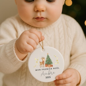 Peut inclure: Ornement de Noël blanc et rond, tenu par un bébé. L'ornement représente un cerf, un sapin de Noël et le texte "MON PREMIER NOËL Ambre 2025" en écriture noire et des étoiles dorées.