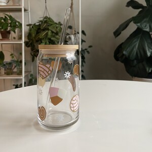 May include: A clear glass jar with a wooden lid and a glass straw. The jar has a variety of colorful designs on it, including hearts, flowers, and swirls.