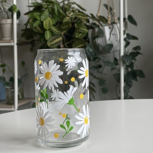 May include: A clear glass drinking glass with a daisy pattern. The glass has white daisies with yellow centers and green leaves. The glass is on a white surface.