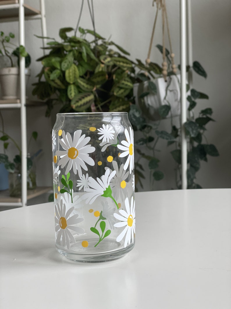 May include: A clear glass drinking glass with a daisy pattern. The glass has white daisies with yellow centers and green leaves. The glass is on a white surface.