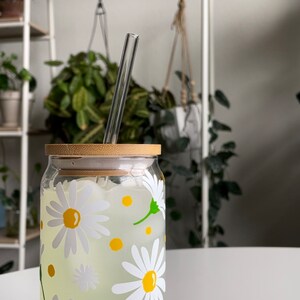 May include: A glass jar with a bamboo lid and a straw, filled with a clear liquid and decorated with white daisies and yellow centers. The jar is sitting on a white surface.