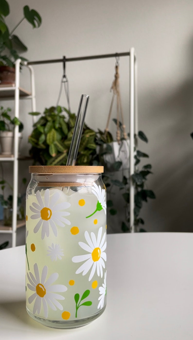 May include: A glass jar with a bamboo lid and a straw, filled with a clear liquid and decorated with white daisies and yellow centers. The jar is sitting on a white surface.