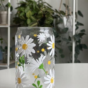 May include: A clear glass drinking glass with a white daisy pattern and yellow accents. The glass has a can-like shape.