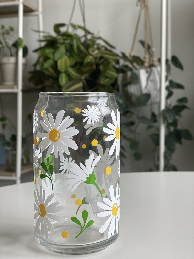 May include: A clear glass drinking glass with a white daisy pattern and yellow accents. The glass has a can-like shape.