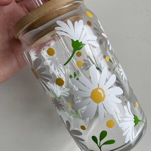 May include: A clear glass reusable water bottle with a bamboo lid and a straw. The bottle has a white daisy pattern with yellow centers and green leaves.