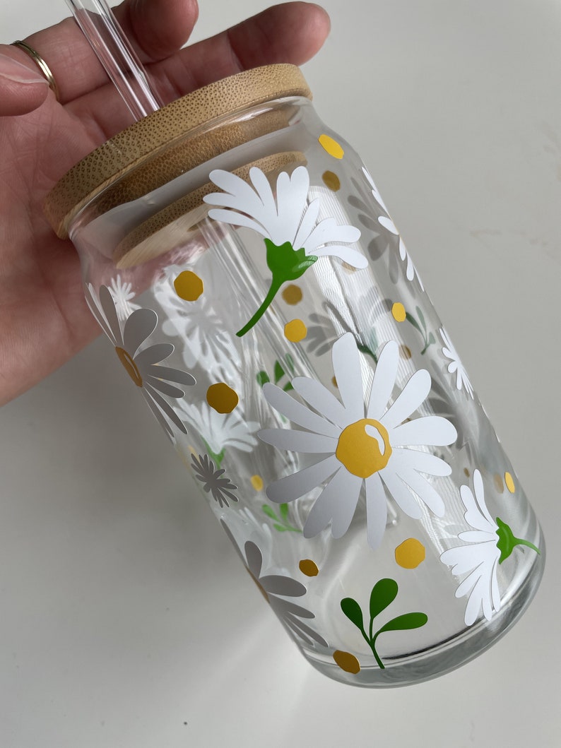May include: A clear glass reusable water bottle with a bamboo lid and a straw. The bottle has a white daisy pattern with yellow centers and green leaves.