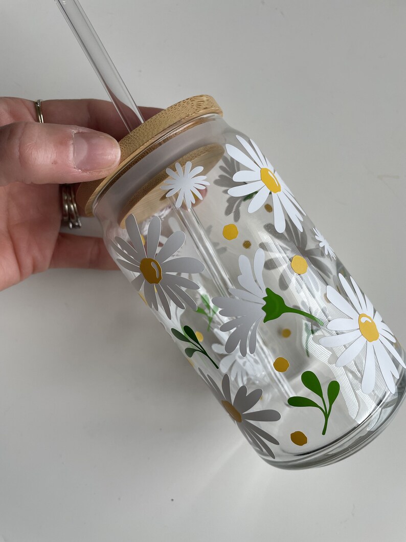 May include: A clear glass reusable water bottle with a bamboo lid and a straw. The bottle is decorated with a white daisy pattern and yellow accents.