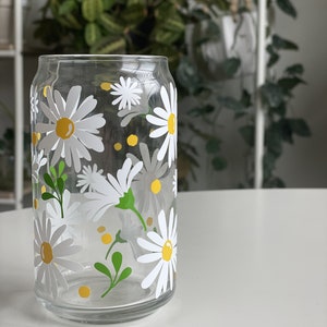 May include: A clear glass drinking glass with a daisy pattern. The glass has white daisies with yellow centers and green leaves.