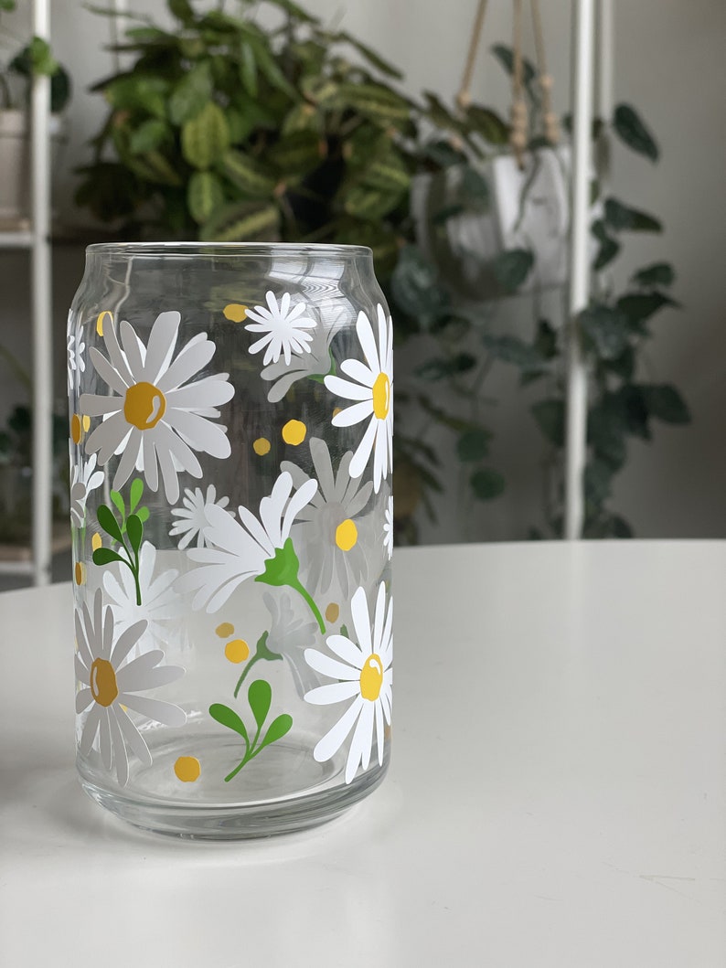 May include: A clear glass drinking glass with a daisy pattern. The glass has white daisies with yellow centers and green leaves.