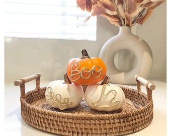 Può includere: Una composizione decorativa a tema autunnale con tre piccole zucche su un vassoio di vimini. Una zucca arancione ha la scritta "BOO" in filo d'oro, mentre le zucche bianche hanno disegni in filo d'oro. Un vaso con foglie secche è sullo sfondo.