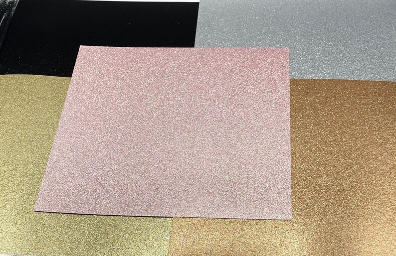 May include: Five sheets of glitter paper in different colors: black, pink, silver, gold, and bronze. The sheets are arranged in a stack, with the black sheet on the bottom and the bronze sheet on the top.