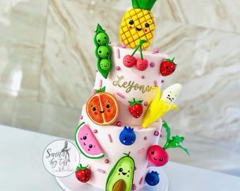 May include: A two-tiered cake decorated with colorful fruit and vegetable characters. The cake is pink with white sprinkles and the name "Leyons" written in gold lettering. The fruit and vegetables include a pineapple, a cherry, a strawberry, a banana, an orange, a watermelon, an avocado, a radish, and peas.