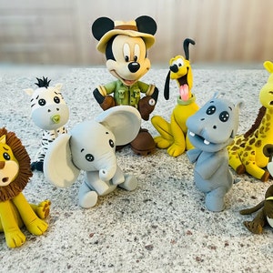 Puede incluir: Un conjunto de nueve figuritas de fondant, que incluyen a Mickey Mouse vestido como explorador de safari, Pluto, una cebra, un elefante, un león, un hipopótamo, una jirafa y un mono. Las figuritas están todas en posición sentada y están hechas para parecer personajes de dibujos animados.