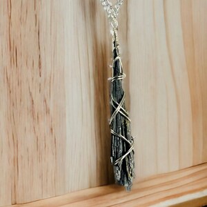 May include: A black kyanite pendant necklace with a silver-colored chain. The kyanite is wrapped in silver-colored wire. The necklace is displayed against a light brown wooden background.