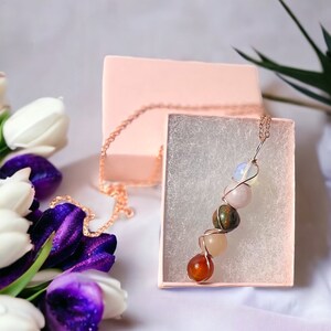 May include: A copper-colored chain necklace with a pendant of various colorful gemstones, including orange, pink, green, and white. The necklace is displayed in a pink gift box with a white interior. White and purple tulips are in the background.