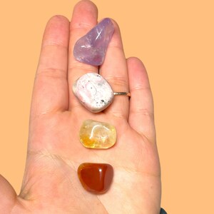 May include: Four tumbled stones in a hand, from top to bottom: a purple amethyst, a pink and white rhodochrosite, a yellow citrine, and a red carnelian.