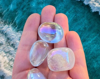 May include: Five clear, polished quartz stones with iridescent, rainbow-like reflections are held in a hand. The stones are oval and rounded, with a smooth, glossy surface. The background shows a turquoise ocean.