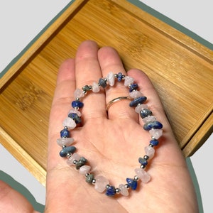 Op de afbeelding: Een armband met rozenkwarts, lapis lazuli en kleine zilveren kralen. De armband bestaat uit onregelmatig gevormde stenen in roze, blauwe en grijze tinten. De armband wordt op een hand getoond.