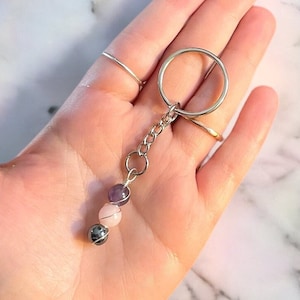 May include: A silver key chain with three gemstone beads. The beads are amethyst, rose quartz, and hematite.