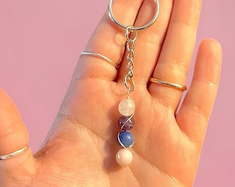 Puede incluir: Un llavero plateado con una cadena y un anillo, adornado con cinco cuentas de piedras preciosas de colores. Las cuentas son rosas, moradas, azules y blancas. El fondo es de color rosa liso.